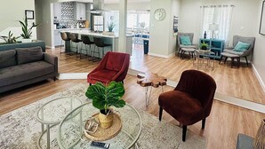 Una sala de estar moderna con una mesa de centro de vidrio, un sillón rojo y un sofá. Se observa una cocina al fondo con un comedor.
