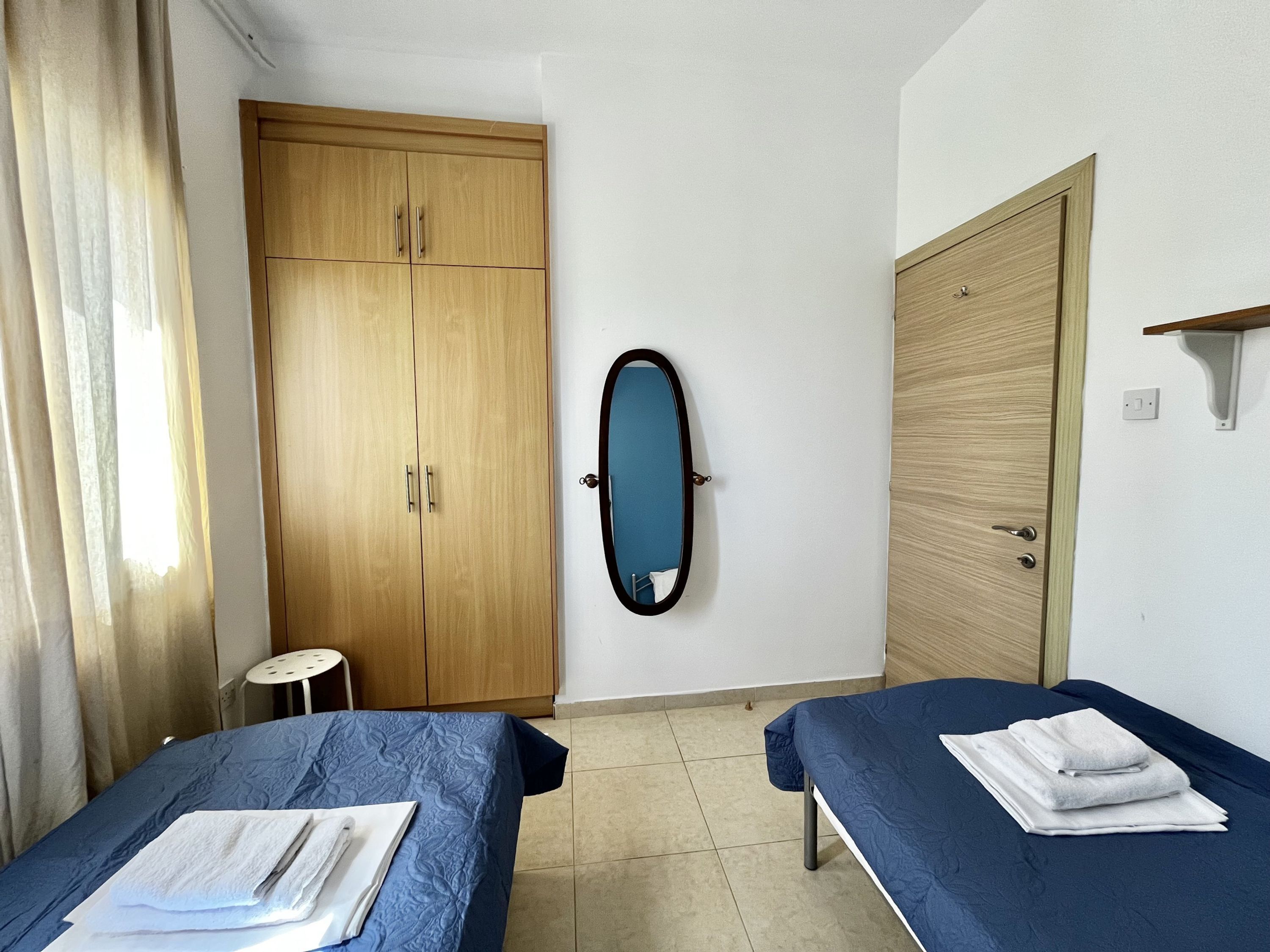Una habitación con dos camas, un armario de madera, un espejo y un taburete pequeño.