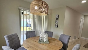 Ein Essbereich mit einem runden Holztisch, um den sechs grau gepolsterte Stühle stehen. Eine gewebte Pendelleuchte hängt über dem Tisch, und an der Wand befinden sich drei gerahmte Bilder.