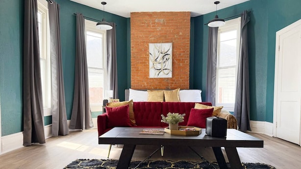A living room with a red sofa, a black coffee table, and a blue rug. There is a brick wall and a window with curtains.