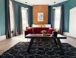 A living room with a red sofa, a black coffee table, and a blue rug. There is a brick wall and a window with curtains.