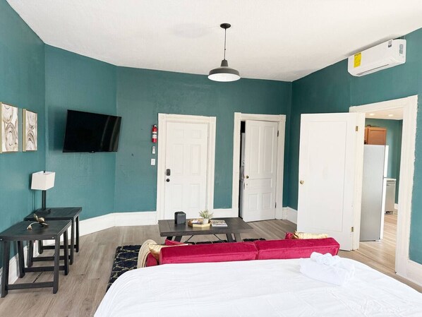 A bedroom with teal walls, a bed, a desk, a TV, and a door leading to a kitchen.