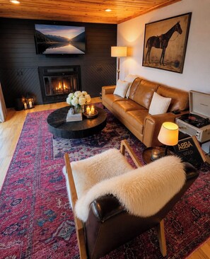 A cozy living room with a leather sofa, a coffee table with candles, a fireplace, and a mounted television. - Boutique Lakefront Cottage, Hot Tub, Sauna (Henderson)