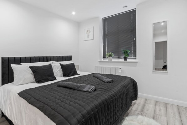 A modern bedroom with a large bed, a window with blinds, a small plant, and a mirror on the wall.
