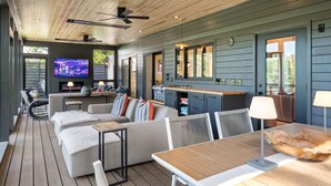 Ein großzügiger Außenbereich mit Holzterrasse, einer Sitzecke mit Sofa und Sesseln, einem Esstisch, einem an der Wand befestigten Fernseher und einer Grillstation.