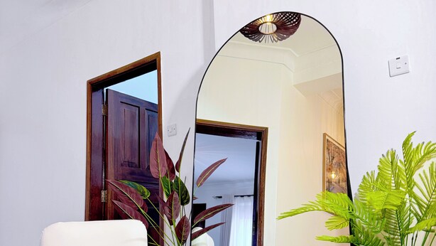 Quarto com um espelho grande, uma cadeira branca, uma planta em vaso e um tapete tecido.