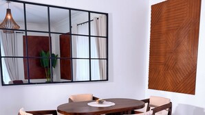 Uma área de refeições com uma mesa de madeira redonda rodeada por seis cadeiras, um espelho grande na parede e um painel de madeira com design geométrico.