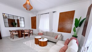 Uma sala de estar moderna com sofá, mesa de centro, mesa de jantar e cadeiras. Há um espelho grande, um vaso de planta e um painel decorativo na parede.