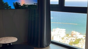 Ein Zimmer mit großem Fenster und Blick auf das Meer und Gebäude, einem flachbildschirmfernseher an der Wand, einem runden Tisch und einer schwarzen Bank.