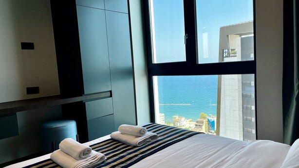 Ein Hotelzimmer mit einem großen Bett, zwei Handtüchern, einer gestreiften Decke, einem dunkel gefärbten Kopfteil und Meerblick durch ein großes Fenster.