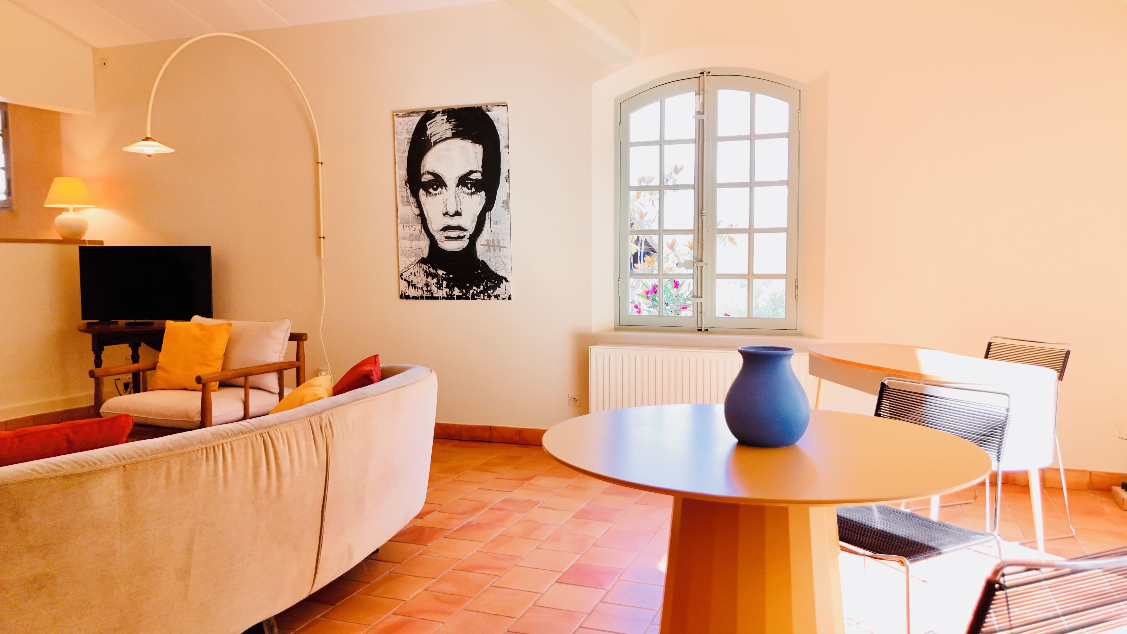 Ein Wohnzimmer mit einer Couch, einem Fernseher, einem Tisch mit einer Vase und einem Fenster mit Blumen.