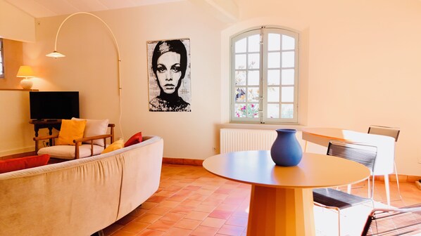 A living room with a sofa, a TV, a table with a vase, and a window with flowers.