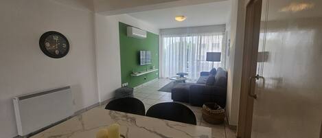 A modern living room with a green accent wall, a flat-screen TV, a sofa, and a dining area with a marble table.