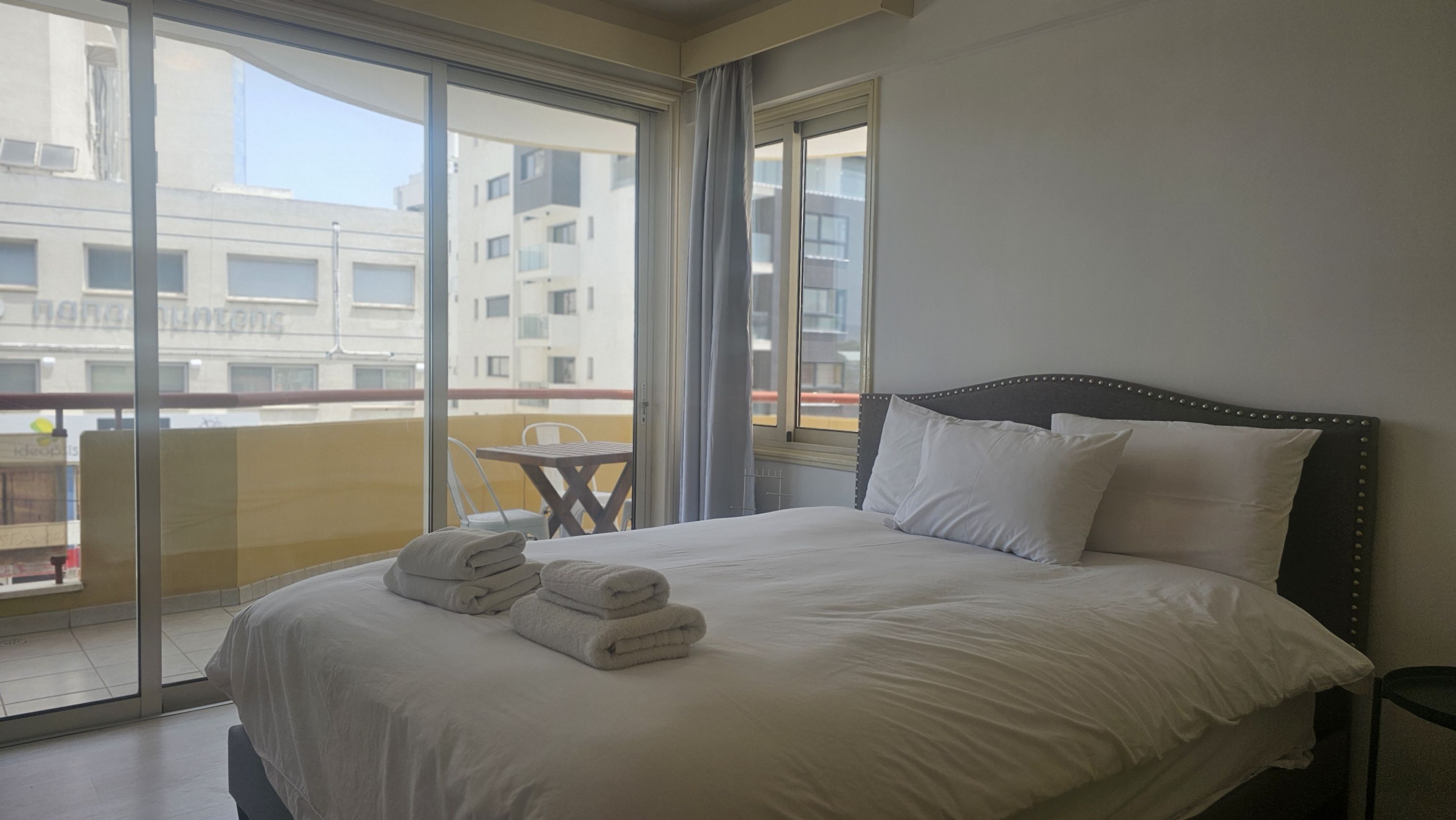 A hotel room with a large bed, two pillows, and a set of folded towels. There is a small table and a chair near the window, which offers a view of the cityscape.