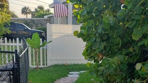 En asfalteret sti fører hen til en hvid bygning med et amerikansk flag, omgivet af frodig vegetation og et hvidt stakittancehegn.