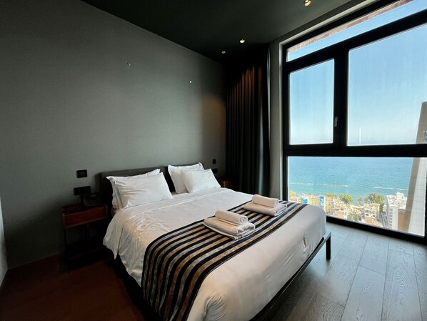A modern hotel room with a large bed, a nightstand, and a view of the city and sea through a large window.