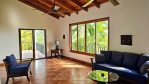 A spacious living room with a wooden ceiling, large windows with a view of greenery, a sofa, a chair, and a small table. - Feel-good oasis Playa Sámara, Ocean view, Private pool, King bed, Dishwasher (Sámara)