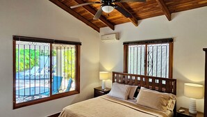 A bedroom with a wooden ceiling, a bed with a headboard, two bedside lamps, and a window with a view of greenery. - Feel-good oasis Playa Sámara, Ocean view, Private pool, King bed, Dishwasher (Sámara)
