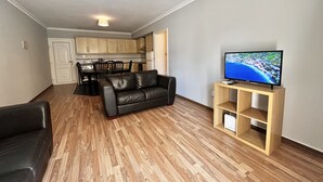 Ein modernes Wohnzimmer mit einer schwarzen Ledersofa, Holzboden, einem Fernseher auf einem Holzschrank und einer Küche im Hintergrund.