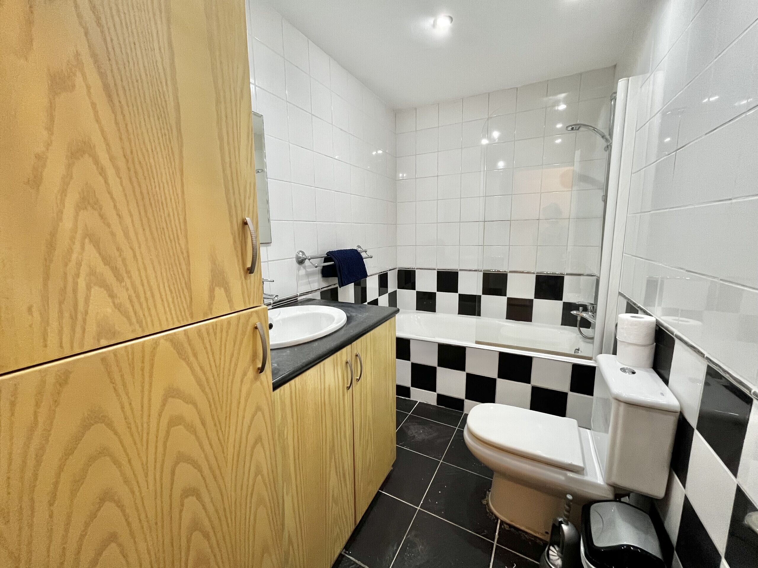 Un baño con tocador de madera, lavamanos blanco, pared a cuadros blanco y negro y un inodoro.