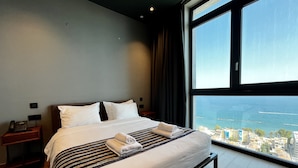 A modern hotel room with a large bed, a nightstand with a phone, and a view of the sea through a large window. - 128 LIM - 2106 Stay Only Tower Suite (Limassol)