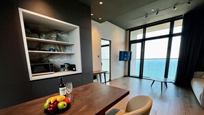 A modern dining area with a wooden table, fruit bowl, and seating. A television is mounted on the wall, and there is a large window with a view of the sea. - 128 LIM - 2106 Stay Only Tower Suite (Limassol)