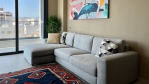 A modern living room with a grey sectional sofa, a colorful abstract painting, a potted plant, and a view of the city through a large window. - STAY Mouson View Apartment (Limassol)