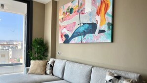 A grey sofa with patterned cushions, a large abstract painting, and a potted plant in a modern living room with a city view. - STAY Mouson View Apartment (Limassol)