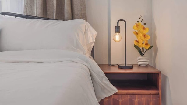 A neatly made bed with white linens, a bedside table with a modern lamp, and a vase with yellow flowers. - Downtown 1 Bedroom Flat with Views (Anchorage)