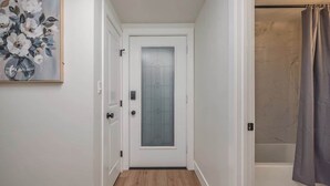 A narrow hallway with a wooden floor, a white door with a frosted glass panel, and a shower area with a grey curtain. - Downtown 1 Bedroom Flat with Views (Anchorage)