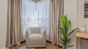 Habitación de esquina con un sillón color beige, una planta en maceta y vistas a través de cortinas translúcidas.