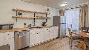 Una cocina moderna con armarios blancos, encimeras de madera, lavavajillas de acero inoxidable y horno empotrado. Hay estantes de madera sobre el lavadero y un comedor con mesa y sillas.