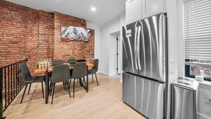 A modern kitchen with a stainless steel refrigerator, a dining area with a brick accent wall, and a window with blinds.