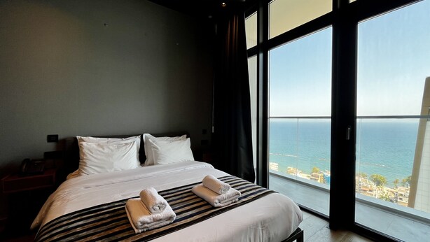 A hotel room with a large bed, two pillows, a bedside table with a phone, and a view of the sea through large windows. - 128 LIM - 2005 Stay Only Tower Suite (Limassol)