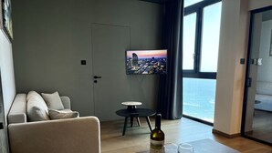 A modern living room with a sofa, a dining table with fruit and wine glasses, a TV on the wall, and a view of the ocean. - 128 LIM - 2005 Stay Only Tower Suite (Limassol)