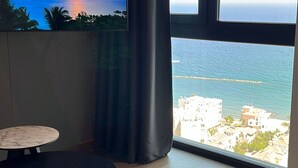 A room with a large window overlooking a coastal view, a television on the wall, and a small round table with a remote control on it. - 128 LIM - 2001 Stay Only Tower Suite (Limassol)