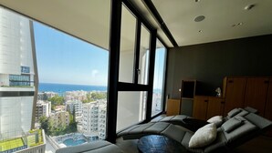 A modern living room with a large window, a sofa, a small table, and a wooden cabinet. - 128 LIM - 2001 Stay Only Tower Suite (Limassol)