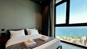 A hotel room with a large bed, two pillows, a striped blanket, and a view of the sea and buildings. - 128 LIM - 2006 Stay Only Tower Suite (Limassol)