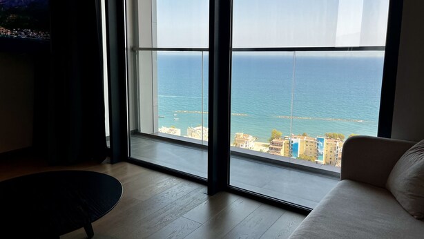 A modern living room with a large window offering a view of the sea and buildings. - 128 LIM - 2006 Stay Only Tower Suite (Limassol)