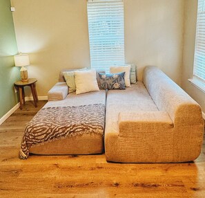 A living room with a large beige sectional sofa, a patterned throw blanket, and several pillows. There is a small wooden side table with a lamp, and a window with blinds. - Modern Downtown 1-Bedroom • Pet-Friendly Stay with Yard (Fort Smith)