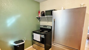Una cocina compacta con un refrigerador de acero inoxidable, un microondas y un horno. Un cubo de basura está colocado contra la pared, y el piso está cubierto de baldosas.