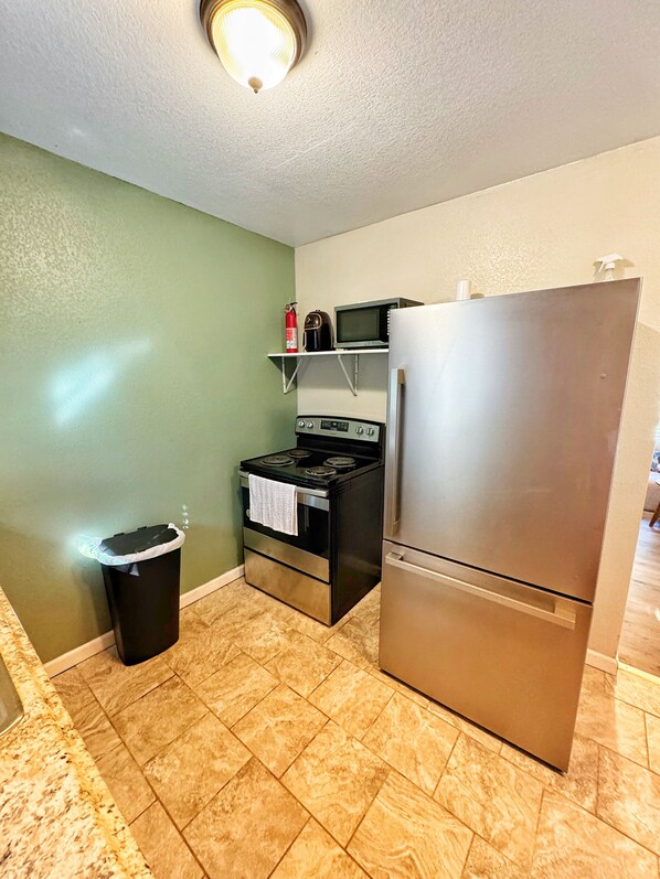 A compact kitchen with a stainless steel refrigerator, a microwave, and an oven. A trash can is placed against the wall, and the floor is tiled. - Modern Downtown 1-Bedroom • Pet-Friendly Stay with Yard (Fort Smith)