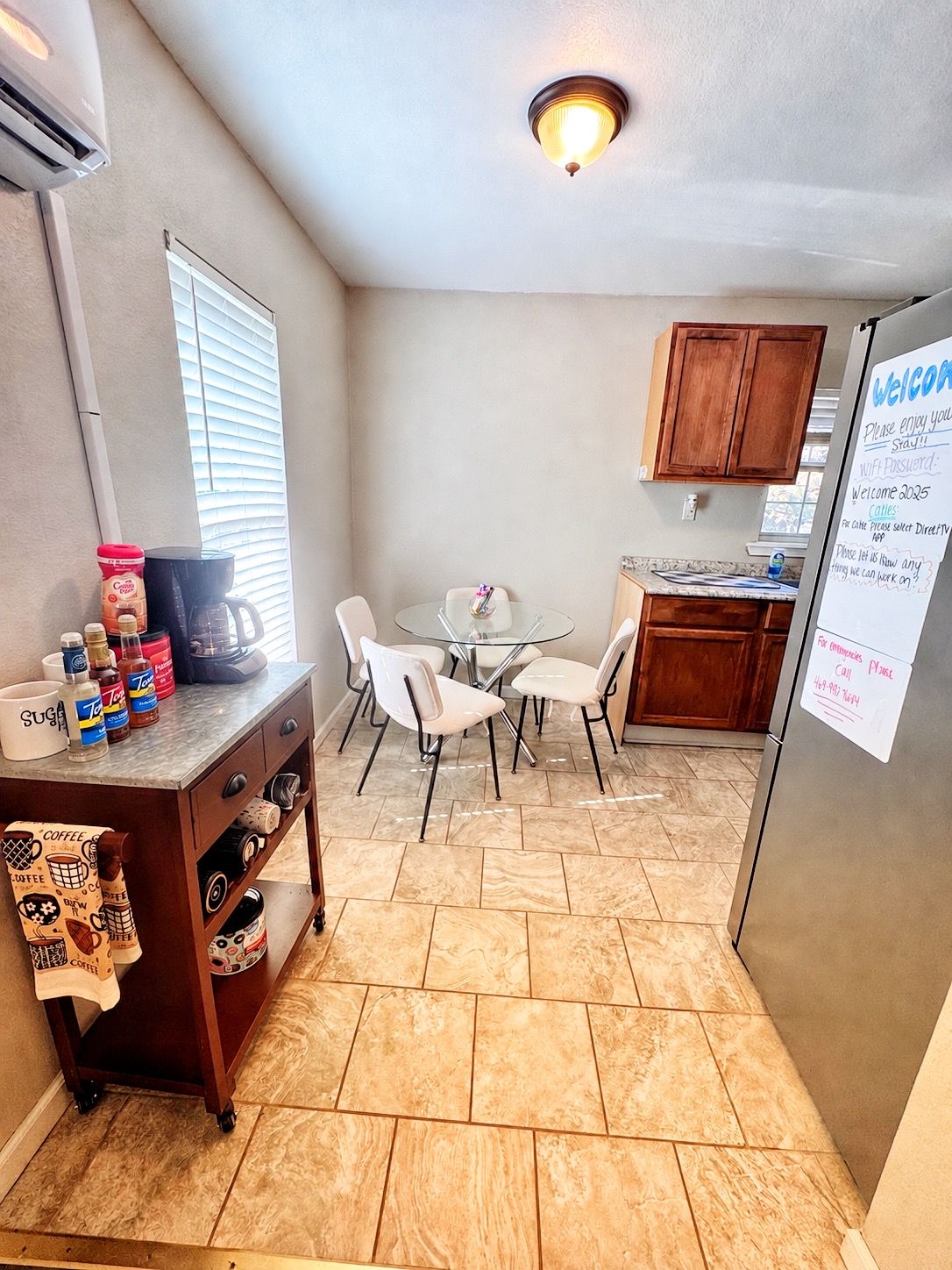 A compact kitchen with a dining area, a coffee station, and a refrigerator with a welcome message.
