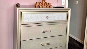 Quarto com televisão de tela plana na parede, placa de madeira com a palavra 'relax' em uma cômoda e uma porta à direita.