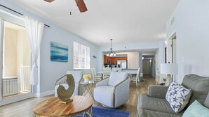 A spacious living room with a grey sofa, a round wooden coffee table, and a blue rug. There is a dining area with a white table and chairs, and a kitchen in the background. - 🦩Sunset Chaser at Ariel Dunes🦩 (Destin)