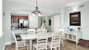 A dining area with a white table and chairs, a chandelier, and a kitchen in the background. - 🦩Sunset Chaser at Ariel Dunes🦩 (Destin)