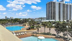 A hotel with a large outdoor swimming pool, a parking area, and a tall building in the background. - 🦩Sunset Chaser at Ariel Dunes🦩 (Destin)
