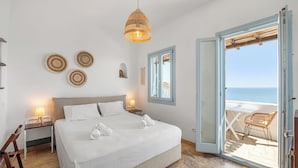 A bedroom with a bed, bedside tables, a hanging light, a view of the sea through an open door, and a chair outside. - Akti Giannaki (Tinos)