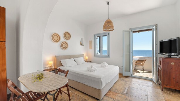 A bedroom with a bed, bedside table, lamp, and a view of the sea. - Akti Giannaki (Tinos)