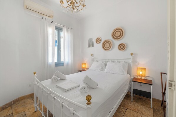 A white four-poster bed with a chandelier, a bedside table with a lamp, and a wall with woven wall art.